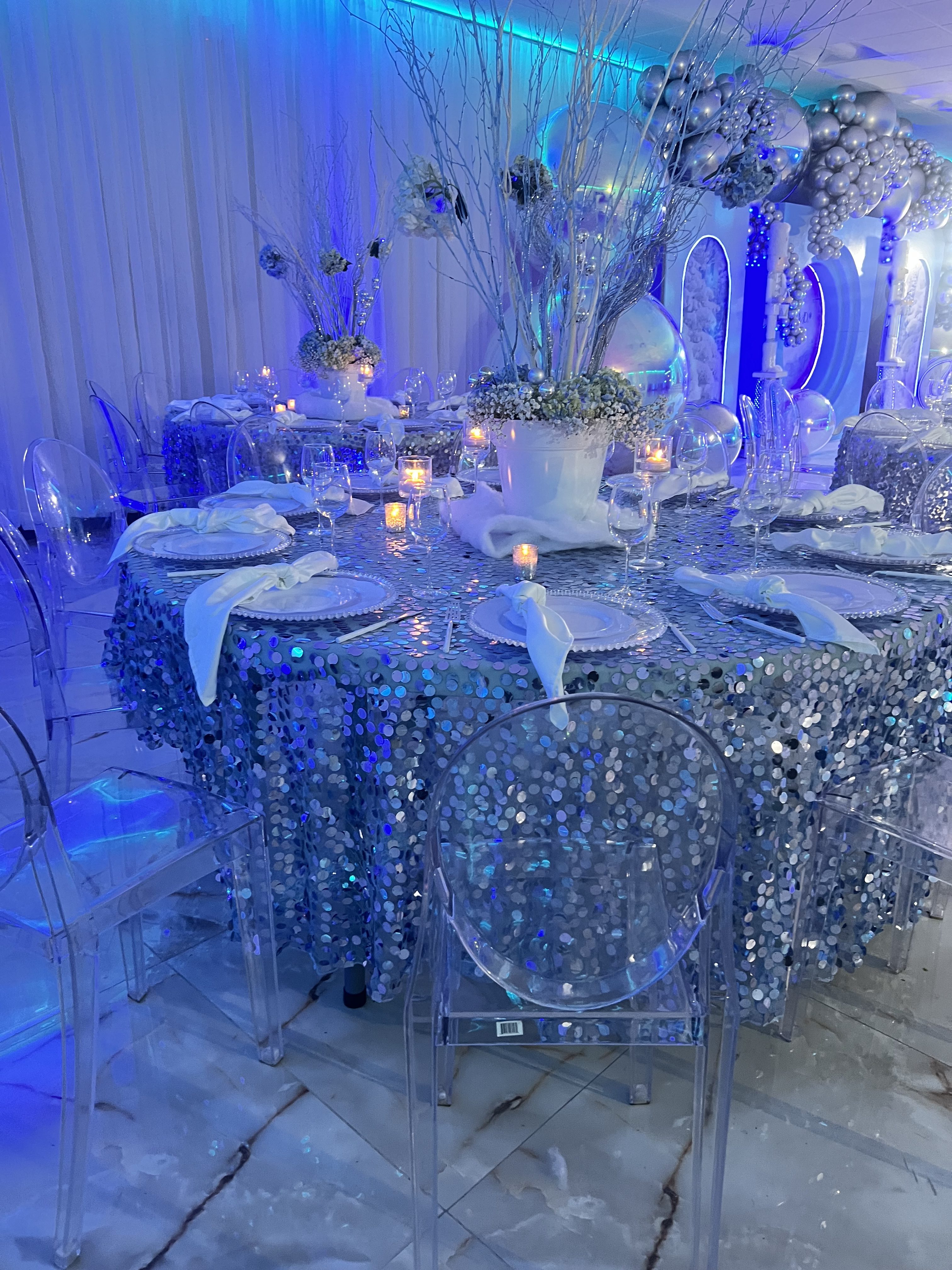A luxurious event table setting with a sparkling blue and silver sequined tablecloth, transparent chairs, and elegant decorations featuring branches, baubles, and candles.
