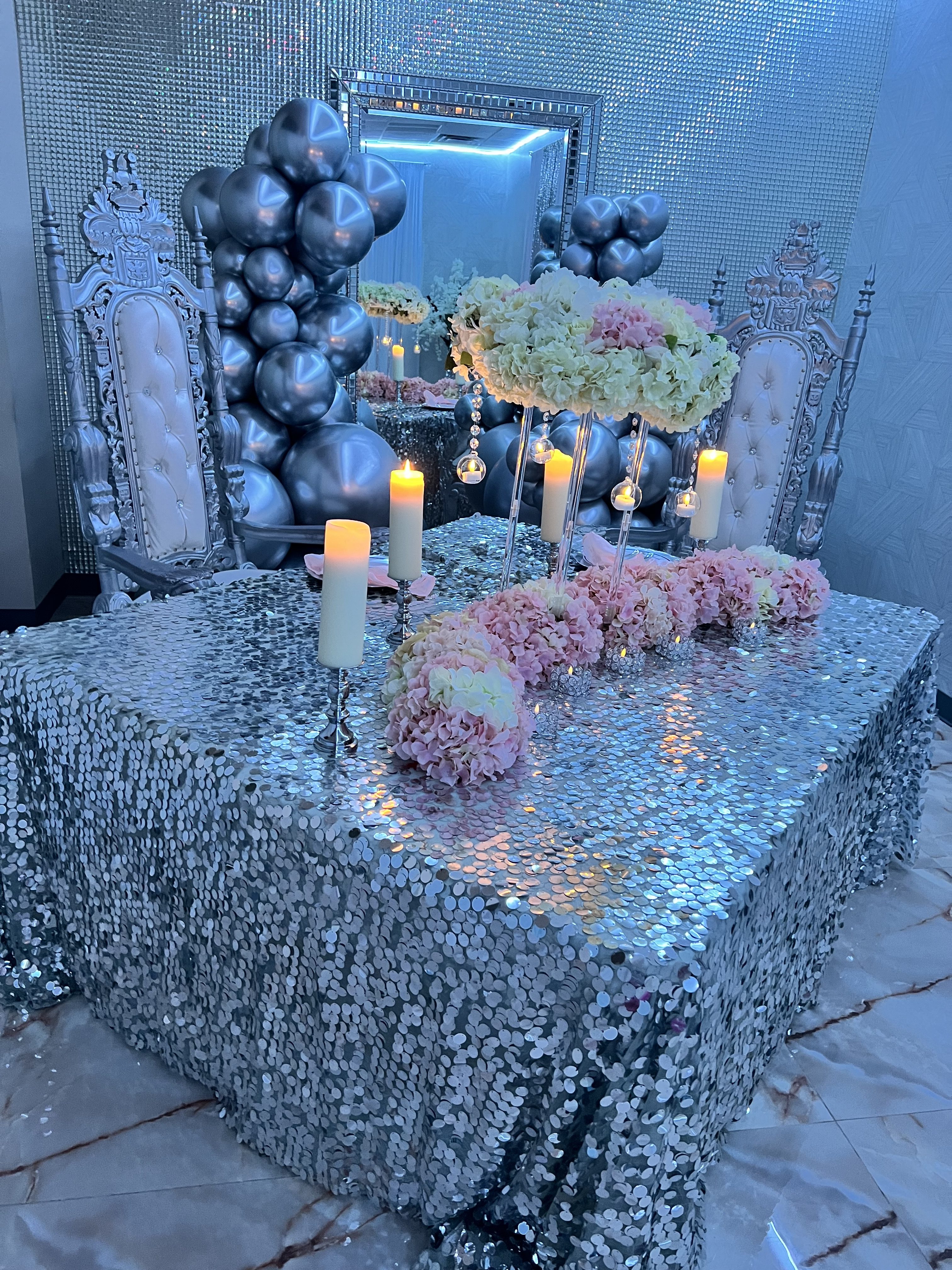 An elegant table setting with a silver sequined tablecloth, surrounded by lit candles, pink and white floral arrangements, and silver-gray balloon decorations.