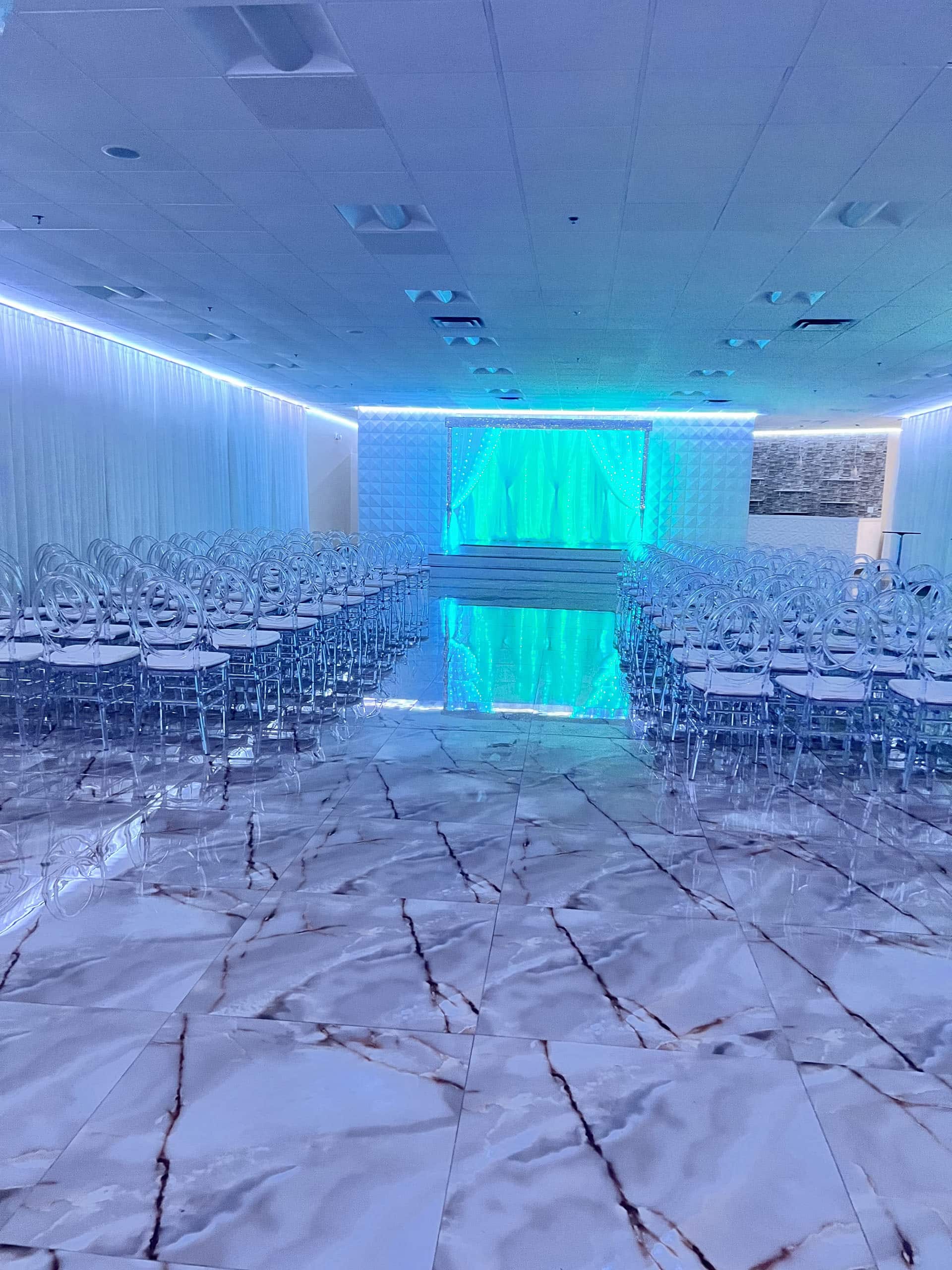 A modern event hall with a marble-patterned floor, rows of clear plastic chairs arranged facing a stage adorned with blue lighting, floor-length white curtains on the walls, and a white ceiling with recessed lighting.